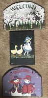Photo showing the three painted slates stacked vertically: top is a white and green tulip welcome slate with paint wear; middle is a black slate with a girl in blue dress and two geese; bottom is a purple slate with two snowwomen dressed in red and purple, holding hands under a red 'WELCOME' sign.