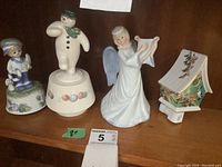 Four ceramic items displayed on a shelf including a little boy figurine, snowman figurine, angel harp player, and birdhouse night light