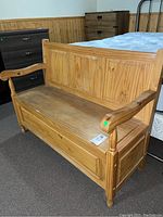 Front view of wooden deacons bench showing armrests, paneled back, and hinged seat, placed on carpeted floor near other furniture.