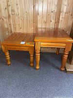 Two wooden end tables with square paneled tops and rounded turned legs, displayed side by side on a carpeted floor against a wooden wall.
