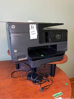 Front and side angle view of the HP Officejet Pro 8610 sitting on a wooden table showing paper tray and connected power cords.