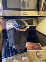 Front and side view of black Keurig coffeemaker on stove with user manual next to it.