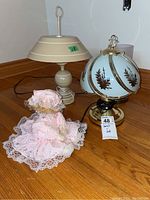 Lot displayed on hardwood floor including the 18 inch beige metal lamp, 11 inch metal base lamp with frosted glass shade and leaf decoration, and a Poter music box doll in pink lace dress and bonnet.