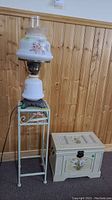 Photo showing full set: white metal plant stand with floral accents, floral hurricane lamp atop the stand, and white felt-lined wooden chest with floral theme beside stand.