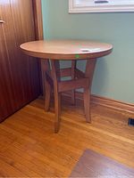 Round wooden table with four legs and smooth natural finish, placed near a wall and window, showing overall condition and style