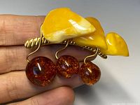 Close-up photo of the brooch held in hand showing the amber beads and leaf pieces with gold-gilt detailing.