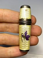 Thread case held in hand showing cream-colored guilloché enamel and purple floral decoration with tarnished sterling silver caps.