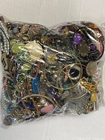 Photo of large sealed plastic bag showing a dense mixture of various jewelry items like bracelets, rings, costume pieces with faux stones and chains tightly packed together.