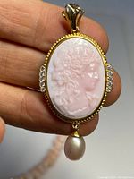 Close-up of oval coral cameo pendant showing detailed carving of woman's profile with gold-gilt frame, rhinestones on sides and dangling pearl.