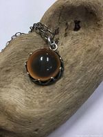 Close-up photo of dome-shaped pendant with brown glass or stone cabochon and twisted sterling silver bezel on a wood background.