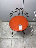 Metal chair with orange round seat and scrollwork backrest shown from front angle on tiled floor.