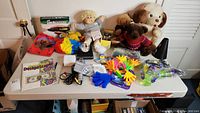 Full view of lot contents spread on white table against white doors, showing doll, bubbles, stuffed animals, noisemakers, game, and coloring book.