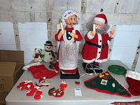 Overview of electric Santa and Mrs. Claus figurines on table with stockings, plush snowman, knitted decorations, and other Christmas decor items