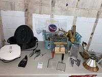 Full view of the lot items laid out on table showing roasting pan with lid, picture lights, basket, grater, kitchen tools, and lamp.