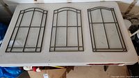 Three glass wall hangings laid flat on a white table, each featuring metal grid frames with arched tops.