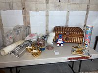 Overview photo showing picnic basket, two swan figurines, stack of plastic 'Grad' cups, tins, plush bear, and rolls of plastic and paper supplies on table against wall.