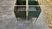 Top angled view of two dark green metal storage containers with four legs each, placed side by side on outdoor pavement. A measuring stick spans across the top measuring approximately 22 inches.