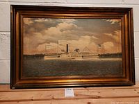 Overall view of framed oil painting showing the Milton Martin boat on water with an American flag and another flag, under a cloudy sky. Wooden frame with brown/gold tones is visible.