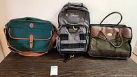 Three bags on a dark surface: a green travel bag, black and gray Indgen backpack, and a green and brown work bag.