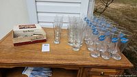 Wooden table with a box of bottle openers, 11 tall clear drink glasses, 4 beer glasses, and 18 Blue Moon pilsner glasses grouped.