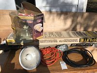 Photo of two hanging shop lights with metal reflectors, orange and black cords, and boxed decorative interior light fixtures on a wooden surface outdoors.