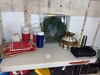 Table with assorted items including stained glass shade, wreath in packaging, thermoses, Coleman water coolers, and boxes