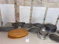 Wide view of the lot showing large oval pewter serving trays, stacks of large and small pewter plates, small pewter handled bowls, and a wooden cutting board on a table against a white wall.