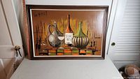 Framed oil on canvas painting showing wine bottles and a glass on a table with a warm brown background.