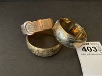 Three gold-tone hinged bangle bracelets arranged on black background, showing floral engraving and buckle style.