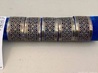 Close-up photo showing three of the six sterling silver napkin rings arranged on a blue fabric holder, highlighting the fine filigree scrollwork and silver borders.