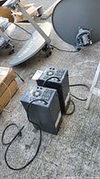 Two black APC battery backup and surge protection units with power cords and fans visible on top, placed on a concrete floor with other equipment around.