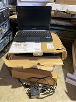 One Chromebook open on its box with power adapter in front and other computer cases in background