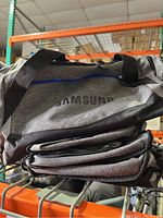 Stacked six Samsung black and silver duffel bags with visible brand logo and shoulder strap on warehouse shelving.