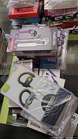 Photo showing a stack of new in box earbuds packaging including Acellories and other brands like Burst Out and Super Bass. Items are assorted and some packages partially visible.