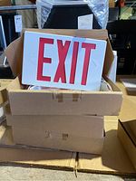 Three cardboard boxes stacked with visible LED exit signs showing red 'EXIT' letters on white faces.
