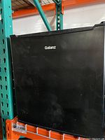 Black Galanz refrigerator freezer unit on warehouse shelf, closed door showing brand logo.