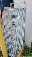 Stack of white metal wire shelves leaning against wall, showing wire grid and round end caps on posts.