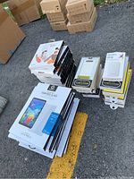 Photo of multiple stacks of boxed wireless stereo headphones, power banks, and LG G Pad 5 tempered glass screen protectors outdoors on pavement.
