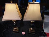 Pair of table lamps shown together on a black cloth background. Lamps have beige square tapered shades and dark wood carved bases, approximately 19 1/2 inches tall.