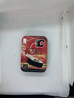 Rectangular metal tin box with Calgary Flames flaming 'C' logo and 'Calgary Flames' text on red flame background. NHL emblem and a hockey puck image also visible on tin.