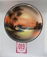 Top-down view of the bowl showing the hand painted river scene with a house, trees, and water under an orange sky.