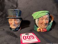 Two small Toby mugs side by side showing faces of an elderly man with top hat and an elderly woman with green headscarf and pipe handle.