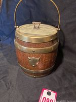 Full view of the vintage tobacco jug showing wooden body with brass bands and handle on a dark fabric background.