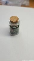 Small glass jar with cork stopper filled with rough green emerald crystals, showing natural and untreated stones.