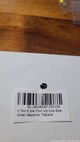 Photo of natural blue-green multicolored oval sapphire placed on paper with label showing carat weight 0.78ct, size 6.2x4.7mm, clarity VS, origin Thailand