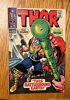 Front cover of The Mighty Thor issue #144 featuring Thor wielding a large green weapon, bright red title and caption 'This Battleground Earth!'
