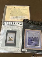 Three sealed cross stitch kits including Lighthouse Classic, Cross My Heart coastal lighthouse, and Early American Sampler Ship Flying Cloud with chart patterns and packaging visible.