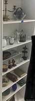 View of a white shelf holding various decorative items including candle holders, glass bowls, wooden decorative basket, ceramic corn dishes, and figurines.