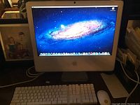 iMac powered on showing macOS desktop with galaxy wallpaper, Apple logo visible on front.