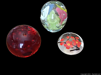 Three art glass paperweights grouped together showing size and color differences: red spherical with bubbles, clear taller with multicolored flowers, small round with red floral design.